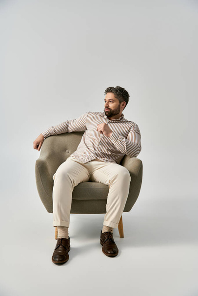 Tyylikäs mies, jolla on parta, istuu tuolissa, syvällä ajatuksissa käsi leualla, harmaata studiotaustaa vasten.. - Valokuva, kuva