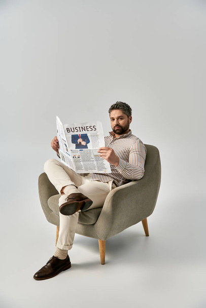 Un hombre elegante y barbudo con un atuendo elegante se sienta en una silla leyendo un periódico sobre un fondo gris del estudio. - Foto, imagen