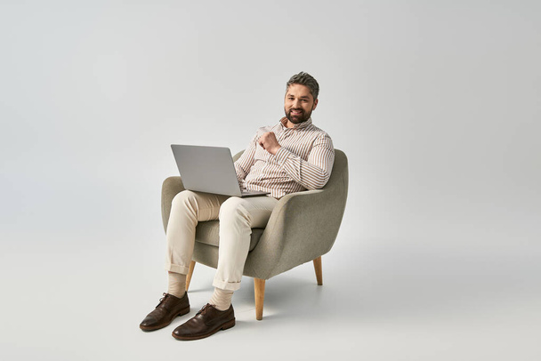 スタイリッシュな服装のひげ付き男は,彼のラップトップに焦点を当てた椅子に座っています. - 写真・画像