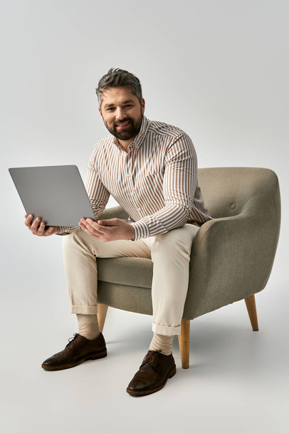 Sakallı şık bir adam bir sandalyeye oturmuş, son moda ve sofistike bir şekilde dizüstü bilgisayarıyla çalışıyor.. - Fotoğraf, Görsel