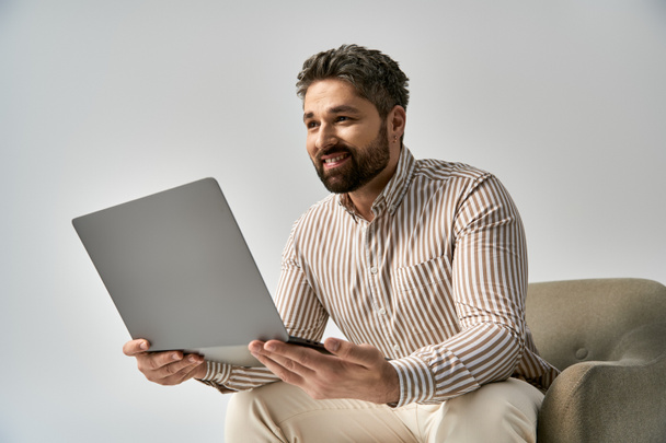 Un homme élégant avec une barbe assise sur une chaise tout en tenant un ordinateur portable dans un cadre de studio sur un fond gris. - Photo, image