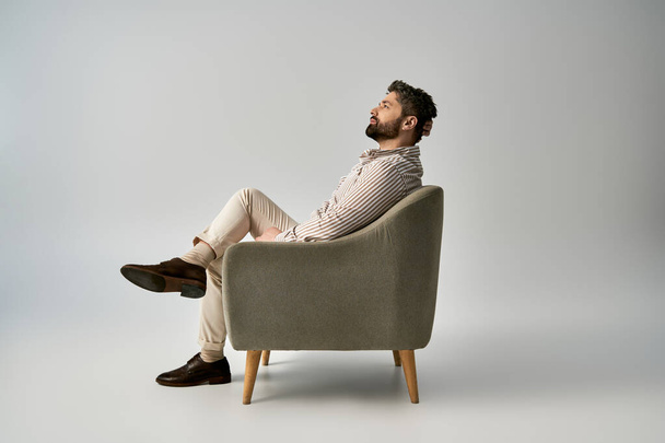 Um homem elegante com barba senta-se em uma cadeira, cruzando as pernas, mostrando elegância e confiança. - Foto, Imagem