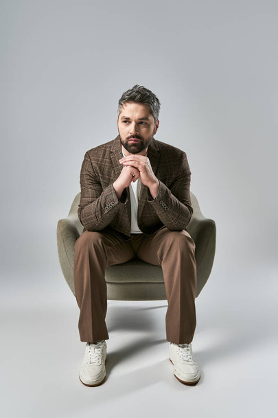 Ein Mann mit Bart sitzt auf einem Stuhl, die Hände in eleganter Pose vor grauem Studiohintergrund gefaltet. - Foto, Bild