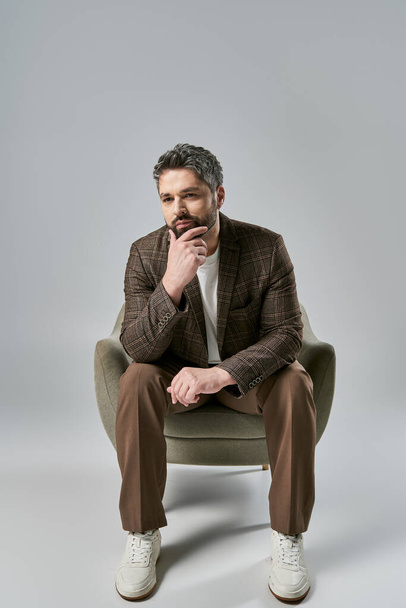 Un hombre barbudo con un atuendo elegante se sienta con la mano en la barbilla, reflexionando profundamente en una pose elegante sobre un fondo gris de estudio. - Foto, imagen