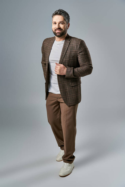 Een boeiende man met een baard slaat een pose in een elegant bruin pak en wit shirt tegen een grijze studio achtergrond. - Foto, afbeelding