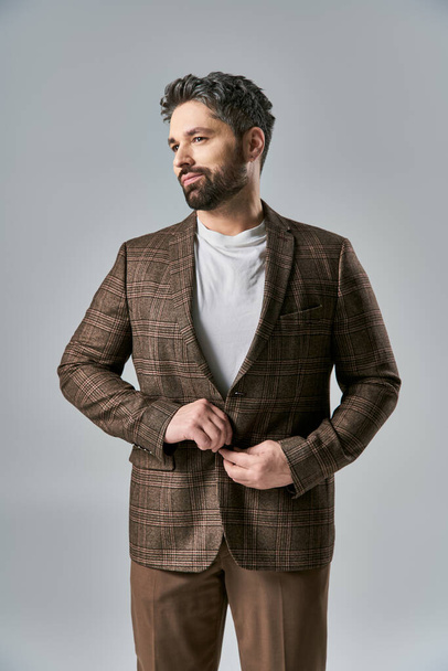 A charismatic man with a beard wearing a brown jacket and pants strikes a pose in an elegant fashion on a grey studio backdrop. - Photo, Image