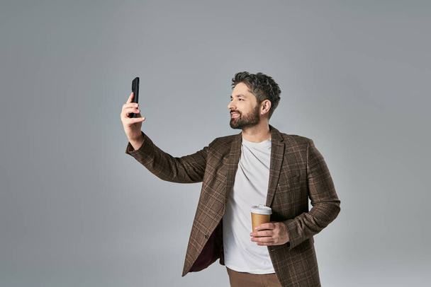 Un homme élégant avec une barbe vêtue d'une tenue élégante prenant un autoportrait avec son téléphone portable sur un fond gris. - Photo, image