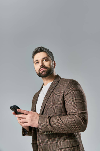 Ein bärtiger Mann im Anzug hält ein Handy in der Hand und strahlt Eleganz in einem Studio vor grauem Hintergrund aus.. - Foto, Bild