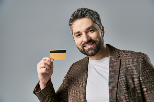 Ein stilvoller Mann mit Bart, der eine Kreditkarte in der Hand hält und vor grauem Studiohintergrund fröhlich lächelt. - Foto, Bild