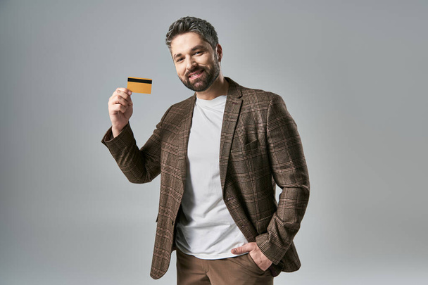 Szakállas férfi elegáns öltözékben magabiztosan tartja a hitelkártyát, bemutatja a modern pénzügyi tranzakciók. - Fotó, kép