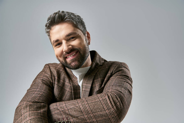 Een stijlvolle man met een baard lacht vol vertrouwen met zijn armen gekruist in elegante kleding tegen een grijze studio achtergrond. - Foto, afbeelding