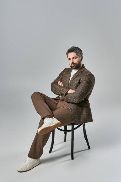 Un uomo barbuto siede con le braccia incrociate, trasudando forza e portamento in abiti eleganti su uno sfondo grigio studio. - Foto, immagini