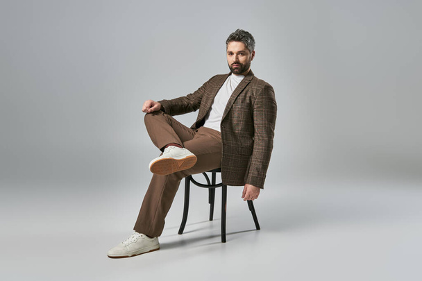 Um homem elegante com uma barba senta-se em uma cadeira, cruzando as pernas elegantemente em uma pose fashion-forward contra um fundo de estúdio cinza. - Foto, Imagem