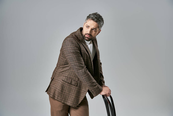 Een bebaarde man die een pose slaat terwijl hij een stoel vasthoudt, gekleed in elegante kleding tegen een grijze studio achtergrond. - Foto, afbeelding