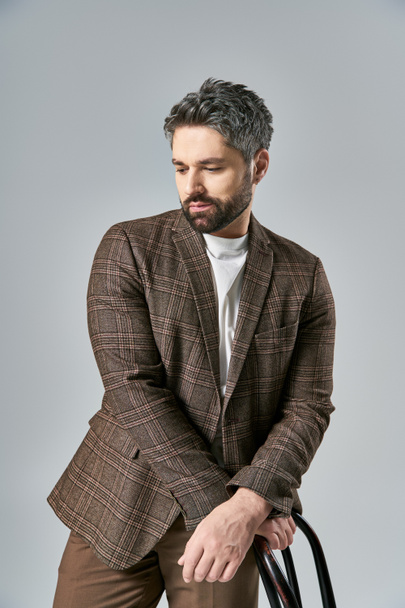 A bearded man in a brown jacket and pants poses with a black chair in a studio setting against a grey background. - Photo, Image