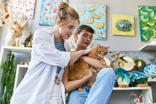 Dos mujeres sosteniendo un gato en sus brazos, rodeadas de artículos de arte en un estudio. - Foto, Imagen