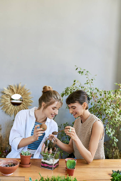 Две женщины, разделяющие момент за столом, окружённые растениями в художественной студии. - Фото, изображение