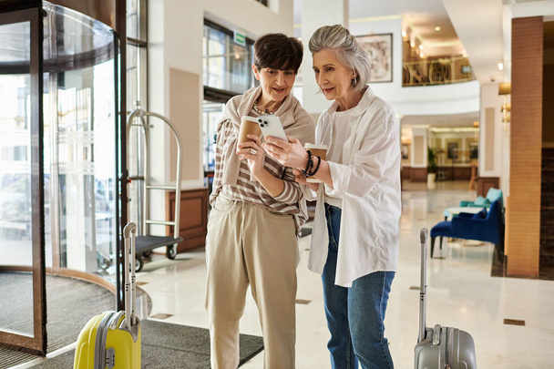 Two women, a loving senior lesbian couple, standing in a lobby engrossed in a cell phone. - Photo, Image