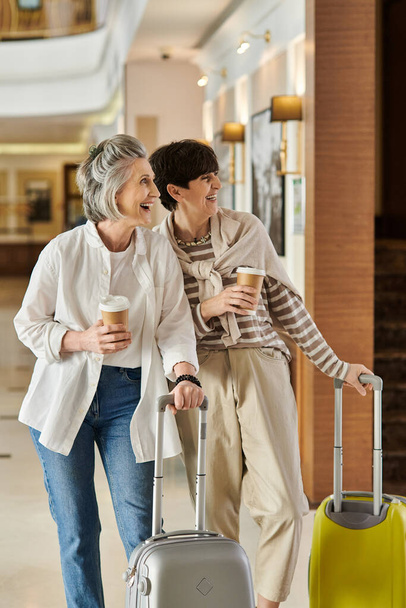 Duas mulheres lésbicas seniores caminham alegremente por um corredor do hotel com bagagem. - Foto, Imagem