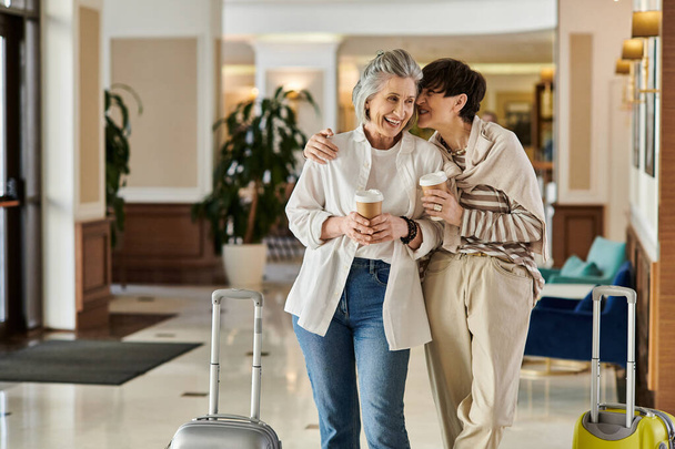 Dos amantes mayores lesbianas de pie juntos en un hotel, compartiendo un momento tierno. - Foto, imagen