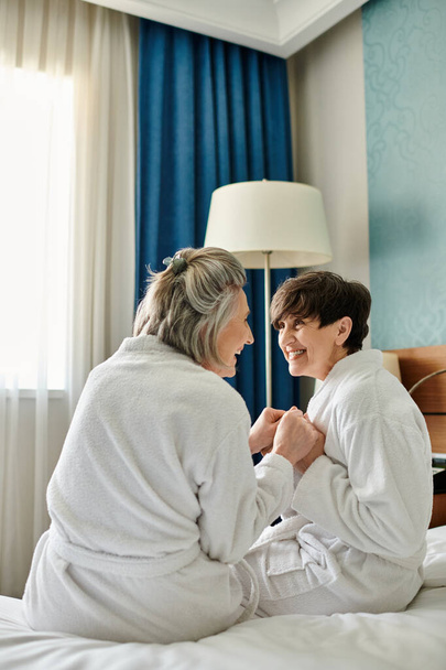 Dwie starsze kobiety, kochająca lesbijska para, siedzą spokojnie na łóżku w pokoju hotelowym. - Zdjęcie, obraz