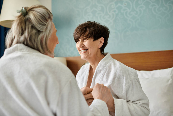 Una mujer con bata de baño sonríe mientras otra mujer se pone su bata en un tierno momento de conexión. - Foto, imagen