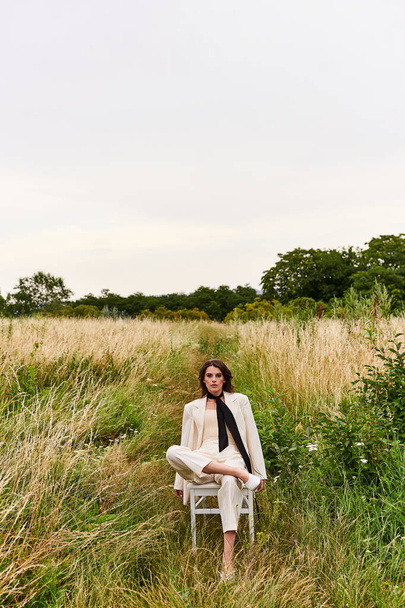 Uma bela jovem mulher de traje branco senta-se em uma cadeira, imersa em serenidade, desfrutando da brisa de verão em meio a um campo cênico. - Foto, Imagem