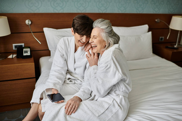 Στοργικό ζευγάρι ηλικιωμένων λεσβιών κάθεται στην κορυφή του κρεβατιού του ξενοδοχείου, μοιράζονται μια τρυφερή στιγμή. - Φωτογραφία, εικόνα