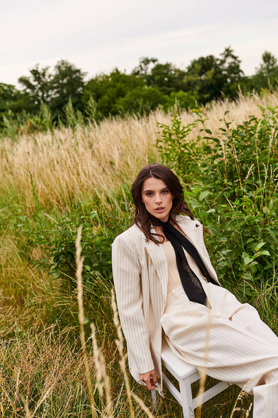 Una hermosa joven vestida de blanco sentada tranquilamente en una silla, empapando la brisa del verano en un exuberante campo. - Foto, imagen
