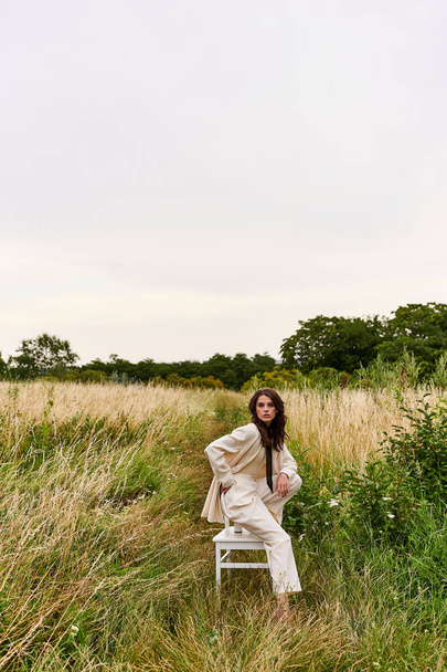 Красива молода жінка в білому вбранні сидить витончено на стільці, насолоджуючись літнім бризом в пишному полі. - Фото, зображення