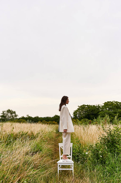 Egy gyönyörű, fehér ruhás fiatal nő áll egy széken, élvezve a nyári szellőt a mezőn.. - Fotó, kép