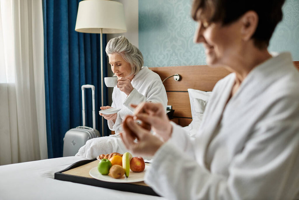 Dos mujeres lesbianas mayores con túnicas blancas, tiernamente sentadas en una cama. - Foto, imagen