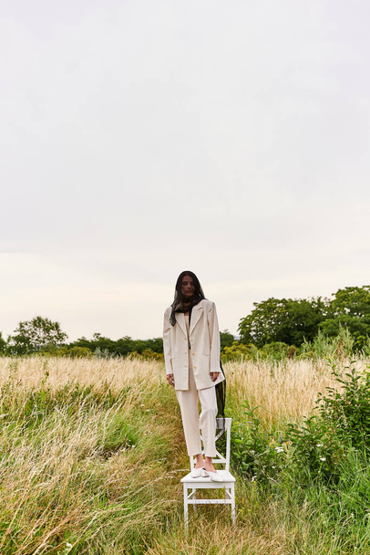 Una hermosa joven vestida de blanco se para en una silla en un campo vibrante, abrazando la brisa del verano. - Foto, imagen