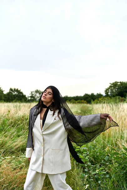 Una hermosa joven vestida de blanco se levanta con gracia en un campo de hierba alta, disfrutando de la brisa del verano. - Foto, Imagen