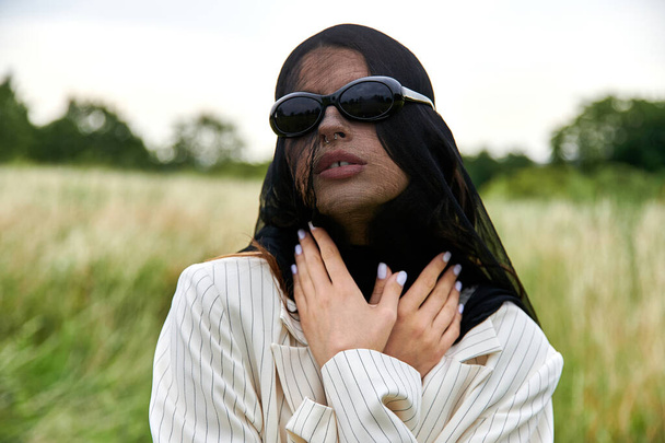 Une belle jeune femme en voile et lunettes de soleil profite de la brise estivale dans un champ de nature. - Photo, image
