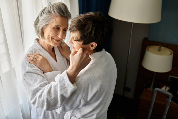 A senior lesbian couple in bathrobes sharing a tender embrace in a hotel. - Photo, Image
