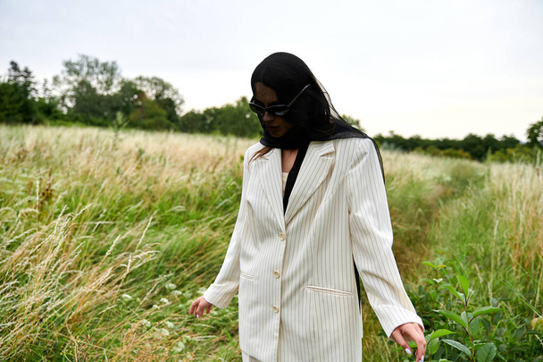 Uma jovem mulher bonita em um terno branco caminha graciosamente através da grama alta em um campo sereno, apreciando a brisa de verão. - Foto, Imagem