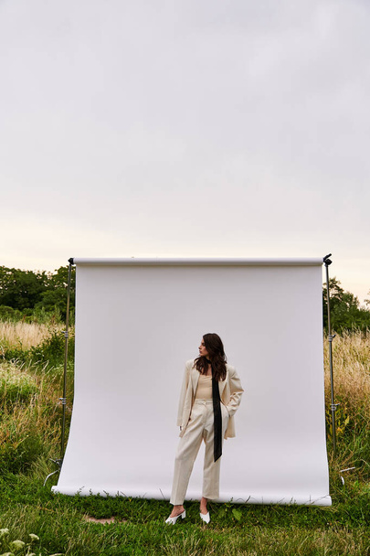Een mooie jonge vrouw in witte kledij staat gracieus voor een witte achtergrond, genietend van de zomerbries. - Foto, afbeelding
