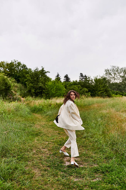 Μια όμορφη νεαρή γυναίκα σε ένα λευκό φόρεμα με χάρη περπάτημα μέσα από ένα καταπράσινο πεδίο, απολαμβάνοντας το καλοκαιρινό αεράκι. - Φωτογραφία, εικόνα