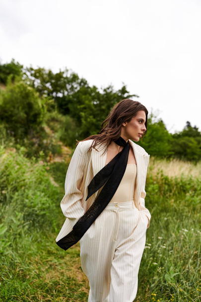 Una hermosa joven con un traje blanco y una bufanda negra disfruta de la brisa del verano en un campo sereno. - Foto, imagen