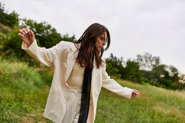 Una hermosa joven con un traje blanco y corbata negra se levanta con gracia en un campo, sintiendo la brisa del verano. - Foto, Imagen
