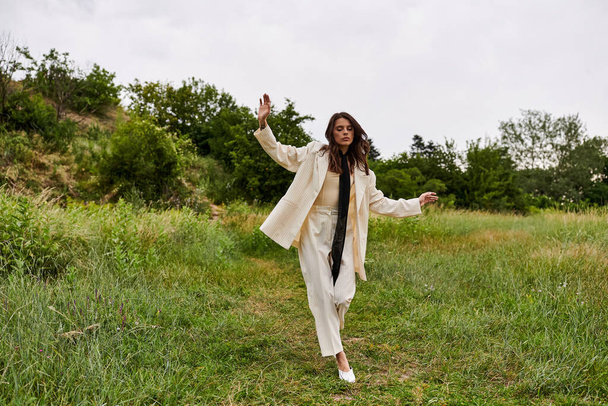 Kaunis nuori nainen valkoisessa takissa kävelee sulavasti rauhallisen kentän läpi nauttien kesätuulesta.. - Valokuva, kuva