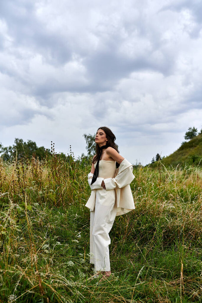 Μια όμορφη νεαρή γυναίκα με λευκή ενδυμασία στέκεται με χάρη σε ένα χωράφι με ψηλό γρασίδι, απολαμβάνοντας το καλοκαιρινό αεράκι. - Φωτογραφία, εικόνα
