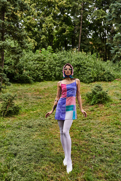 Piękna młoda kobieta w kolorowej sukience i okularach przeciwsłonecznych stoi z wdziękiem na polu, ciesząc się letnią bryzą. - Zdjęcie, obraz