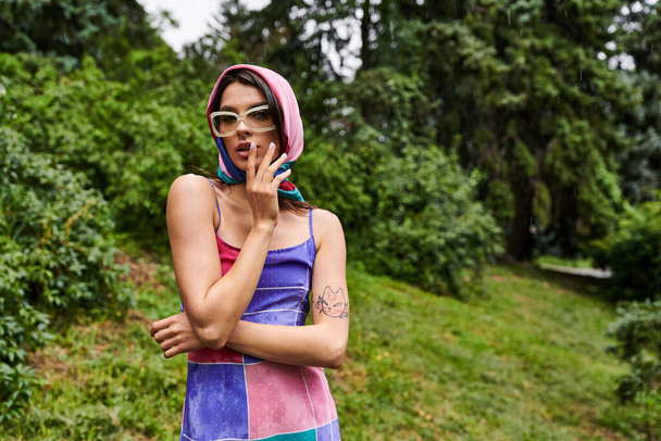 junge Frau genießt die sommerliche Brise in der Natur, trägt ein lebendiges Kleid und eine stylische Sonnenbrille. - Foto, Bild