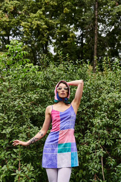 A stunning woman in a vibrant dress and sunglasses stands gracefully in a picturesque field, savoring the gentle summer breeze. - Photo, Image