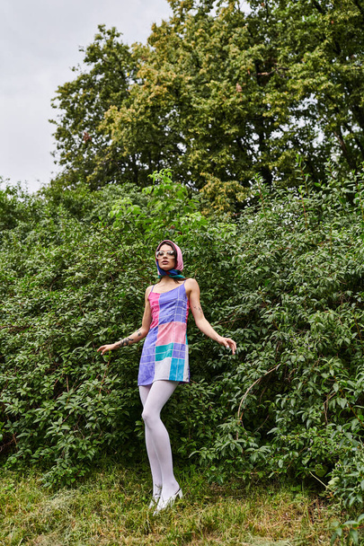 Μια νεαρή γυναίκα με ένα ζωντανό φόρεμα και γυαλιά ηλίου στέκεται σε ένα χωράφι με δέντρα στο παρασκήνιο, μούσκεμα στο καλοκαιρινό αεράκι. - Φωτογραφία, εικόνα