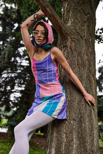 Красива молода жінка в барвистій сукні спирається на дерево, заспокоюючи літній бриз і спокій. - Фото, зображення