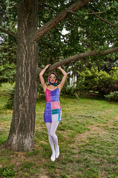 Μια όμορφη νεαρή γυναίκα με ένα ζωντανό φόρεμα και γυαλιά ηλίου στέκεται δίπλα σε ένα δέντρο, απολαμβάνοντας το καλοκαιρινό αεράκι σε ένα ήρεμο πεδίο. - Φωτογραφία, εικόνα