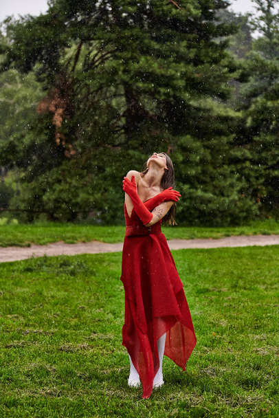 Egy elbűvölő fiatal nő élénk piros ruhában, kecsesen áll az esőben, körülölelve az elemeket higgadtan és elegánsan.. - Fotó, kép